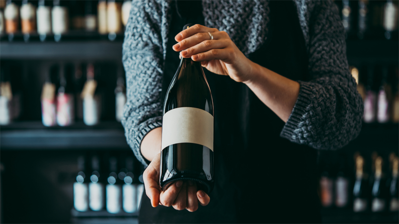 Osoba ukazuje lahev vína. Etiketa vína je prázdná.