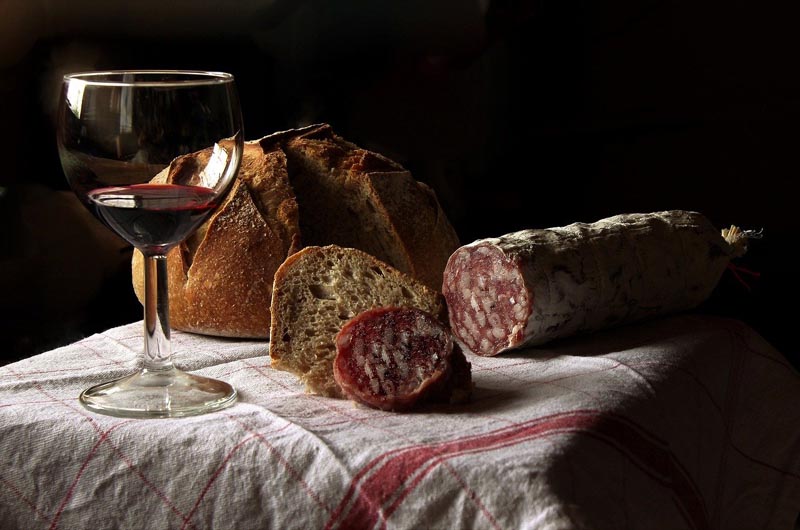 sklenka červeného vína stojící na stole vedle nadýchaného bochníku chleba a klobásy